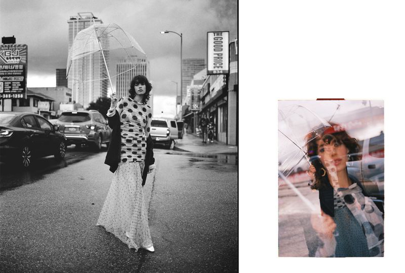 A model stands on a wet street in black and white holding an umbrella, wearing a dotted top and long skirt, with cars and buildings.