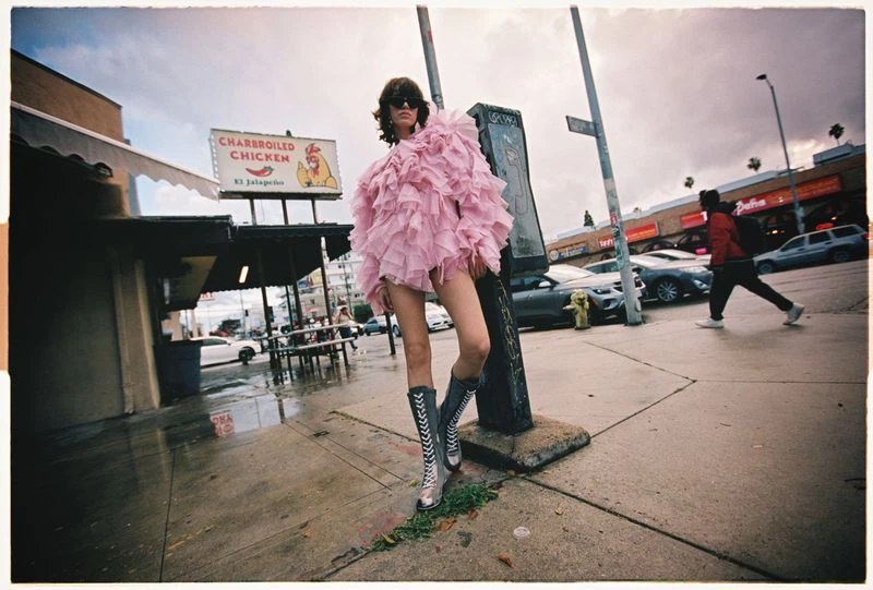 A fashion model in a pink ruffled dress poses on a city sidewalk next to a street sign and a restaurant with a 'Charbroiled Chicken' sign.