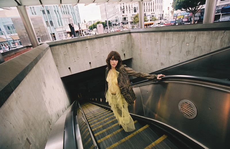 Woman in yellow dress and brown jacket poses on escalator inside a subway station with buildings in the background in an eye-level shot.