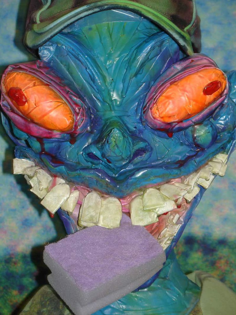 A painted monster mask with teal skin, orange eyes, and a strange expression sits with a purple sponge near its mouth in a close-up shot.