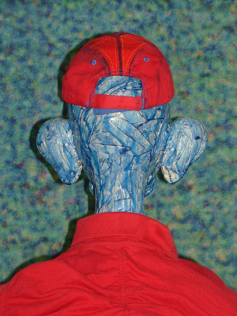 Sculptural figure from behind wearing red baseball cap and red shirt against speckled blue-green background, crafted of plastic wrap.