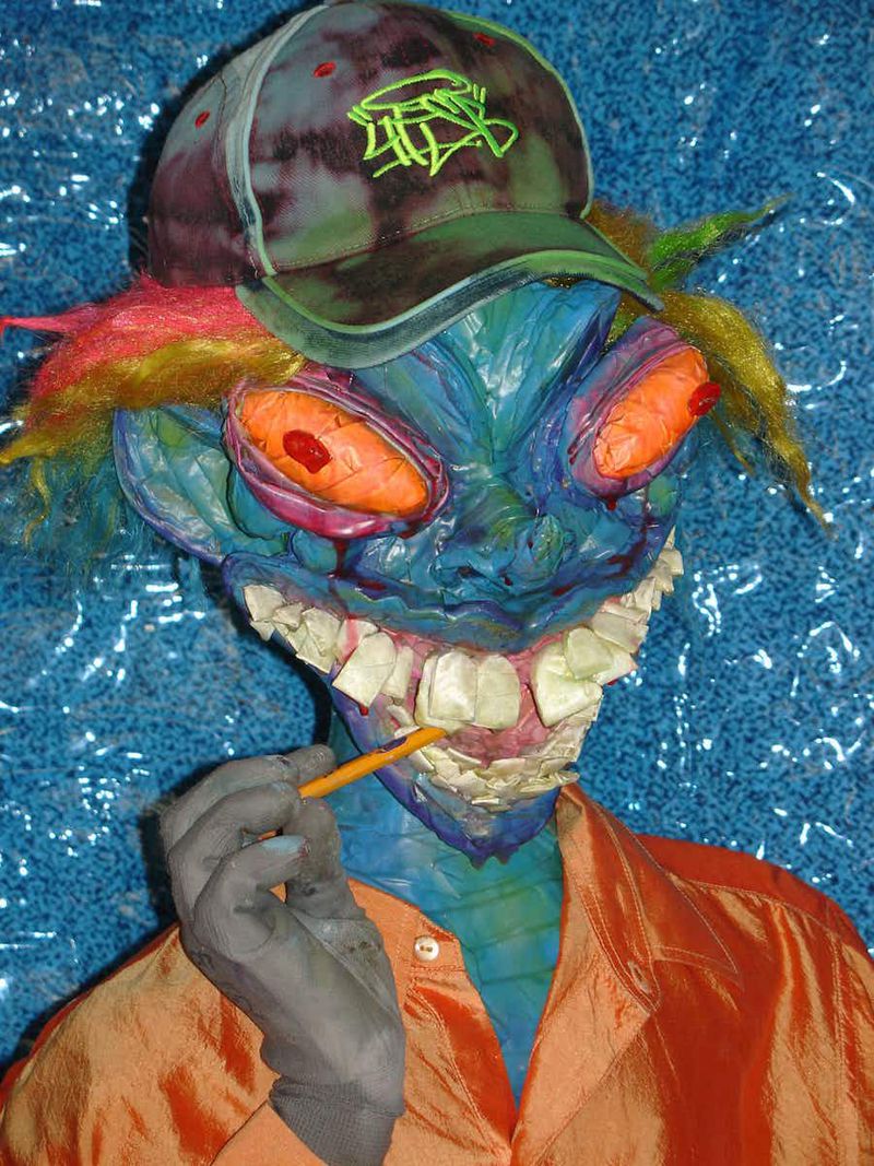 Artistic sculpture of a blue-skinned person with large orange eyes, wearing a colorful hat and an orange shirt, brushing their teeth.