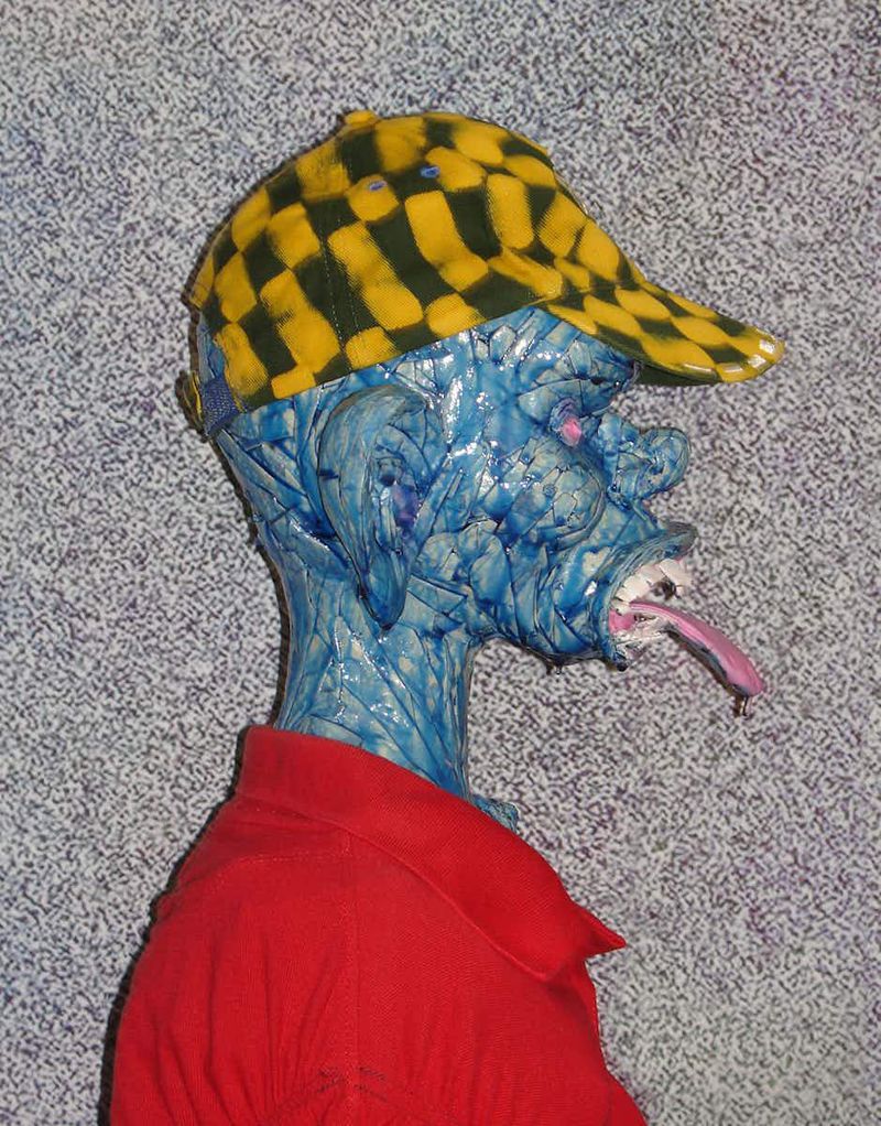 Profile shot of a blue, sculpture-like man with a red shirt and a yellow checkered cap sticking his tongue out.