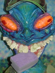 Close-up of a sculptural blue head with large orange eyes and white teeth biting a purple sponge for The Face Magazine