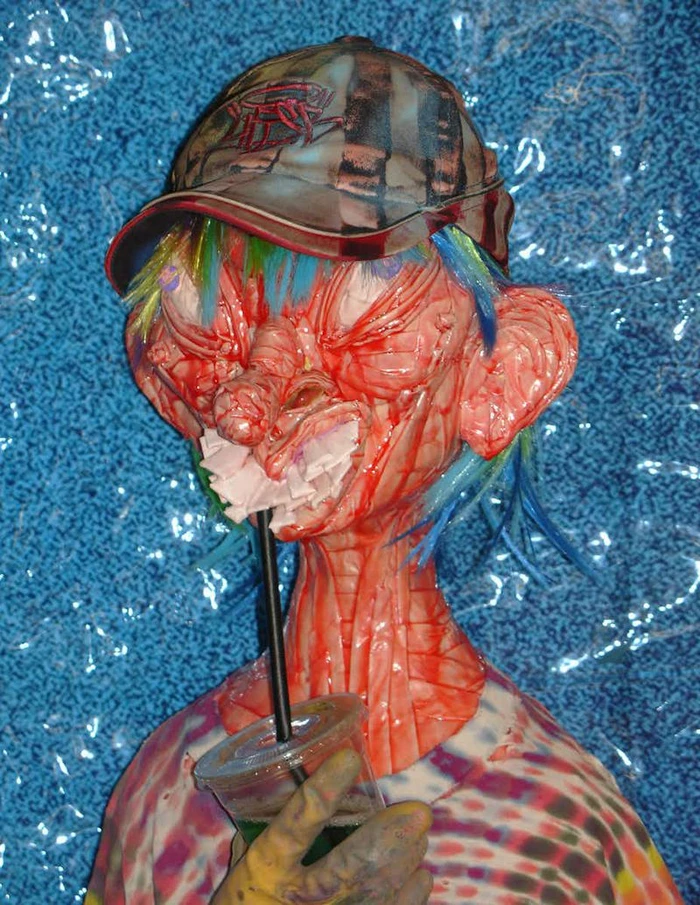 Close-up portrait of a surreal sculpture with textured red flesh-like skin, a baseball cap, and a drink with a straw