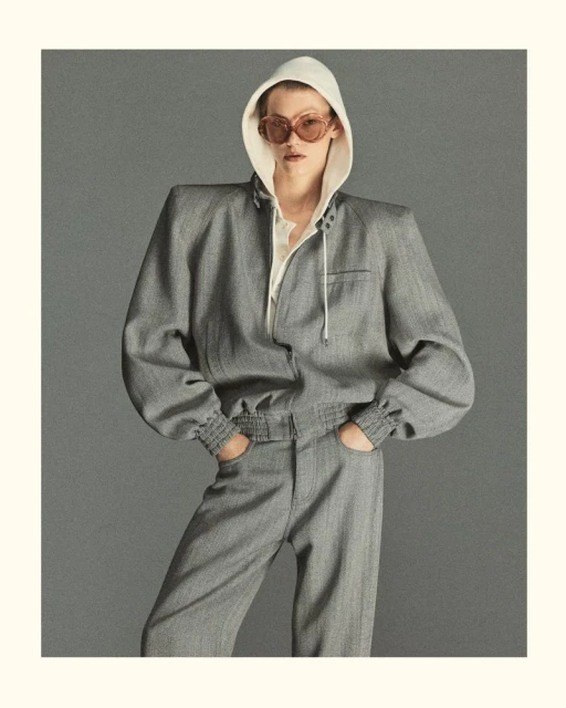 Adult female fashion model poses in gray hooded jacket and pants in a studio setting with neutral background, wearing oval sunglasses.