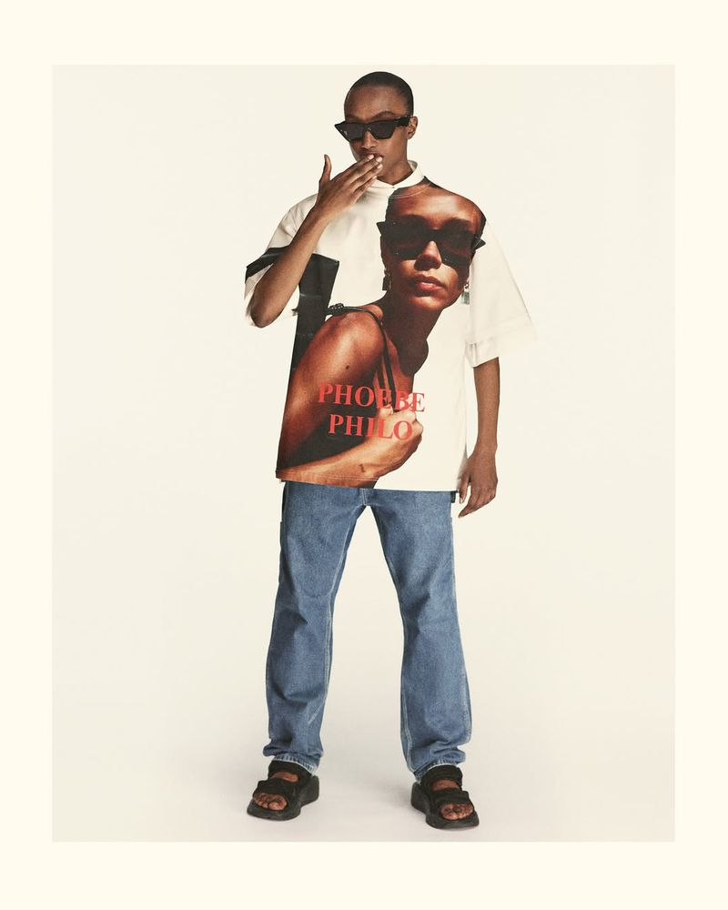 A person in jeans and sandals stands in a studio. They wear a white T-shirt printed with an image and lettering. Sunglasses frame the face.