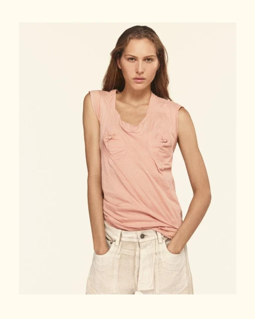 Portrait of a young woman in a pink tank top and off-white shorts looking directly at the camera with hands in pockets.