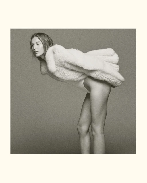 Studio shot of an adult woman wearing a white fur coat and bodysuit, bending forward in a neutral studio setting with a neutral background.