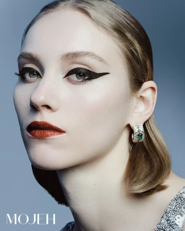 Close-up beauty shot of a fair-skinned woman with dramatic black winged eyeliner, dark red lips, statement earrings, and a sequined dress.