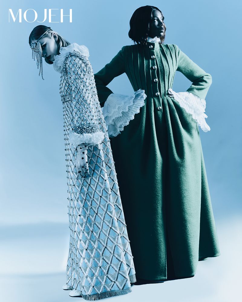 Two women pose against a pale-blue backdrop, modeling a light-blue beaded gown and a green coat with white frilled cuffs. Fashion.