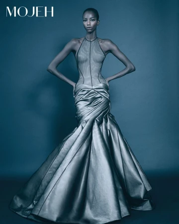 Elegant woman in a silver, sleeveless gown with a trumpet skirt, hands on hips, posing against a solid blue background in studio lighting.