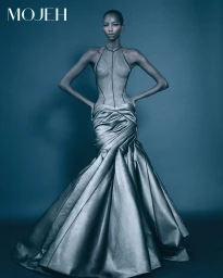 Model in a halter-neck gown with a sheer bodice and voluminous metallic skirt standing against a dark blue studio background