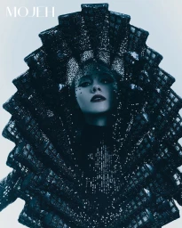 Model wearing an ornate sculptural black embellished headpiece against a plain light background