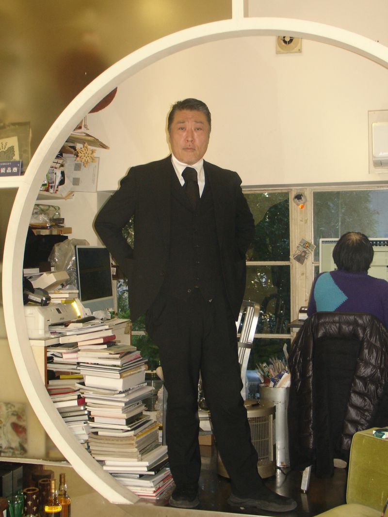 Asian adult man in a black suit, standing in a room with circular frame, a stack of books, and another person working in the background.