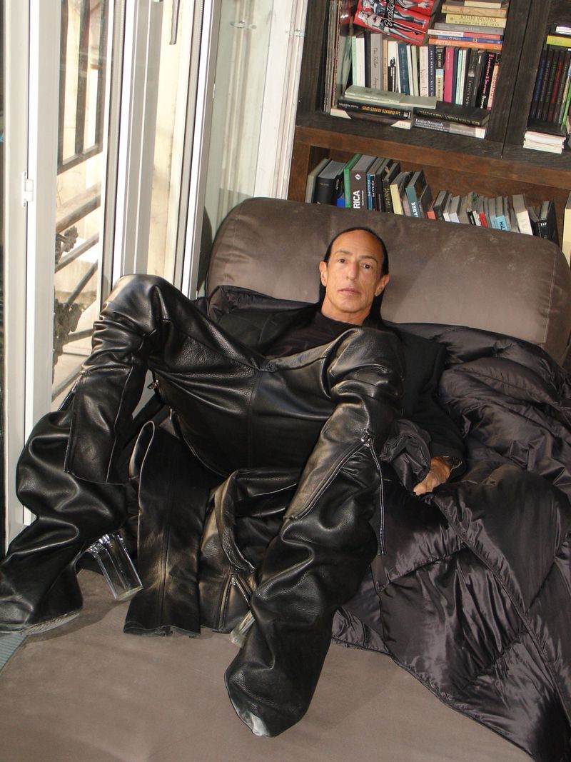 Man in black reclining in a dark chair, wearing oversized leather trousers in an indoor setting with a bookshelf in the background.