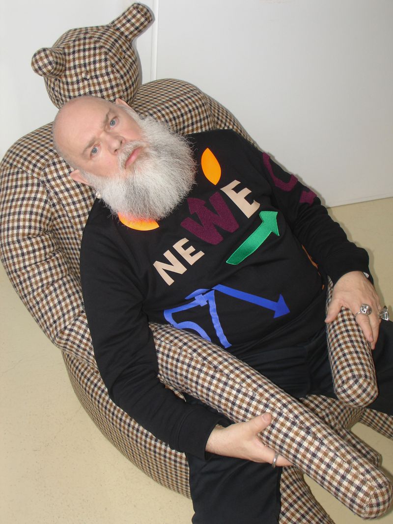 Bald man with white beard sits on a teddy bear chair. He wears a black sweater with colorful letters and shapes. Indoor shot.