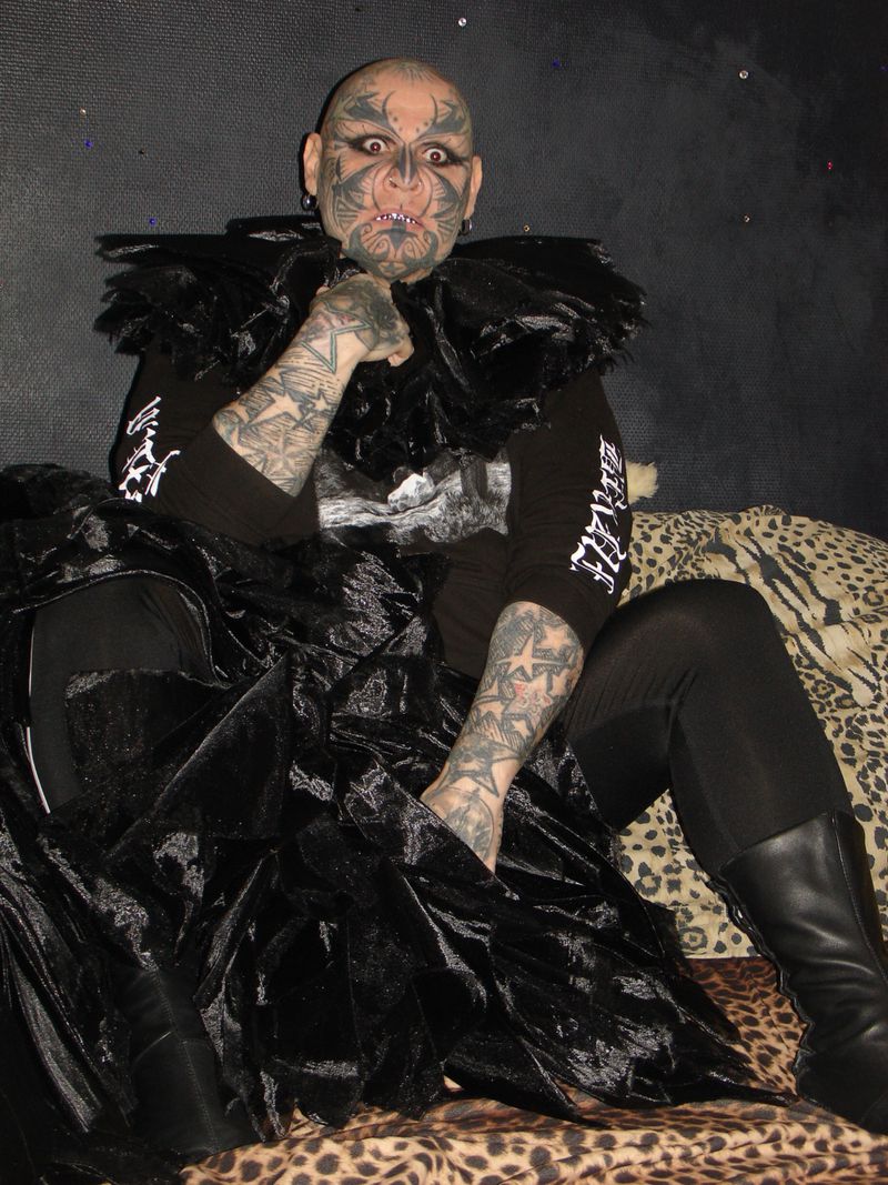 Person with face tattoos seated on a leopard print cushion wearing a black outfit with ruffled collar and black platform boots.