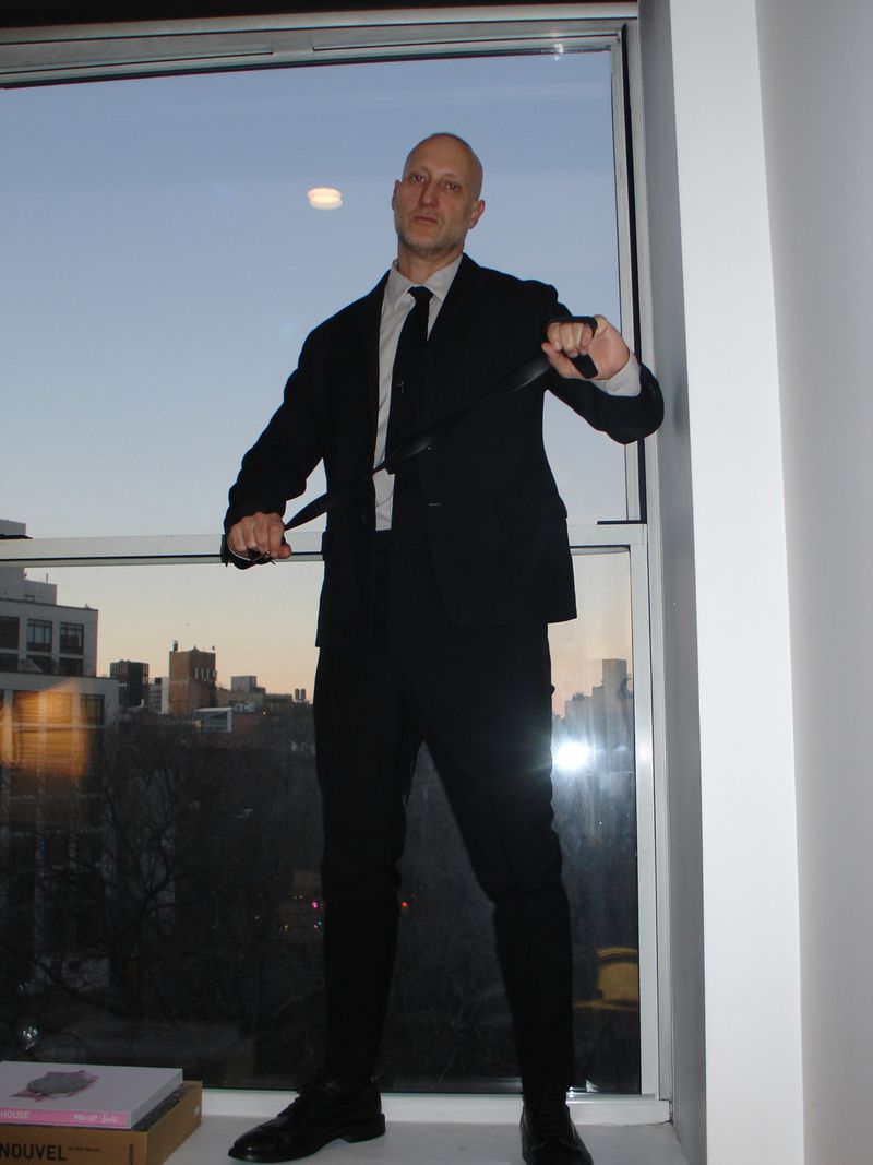 Bald adult male in a dark suit poses indoors by a window at dusk, holding knives, with city buildings in the background.