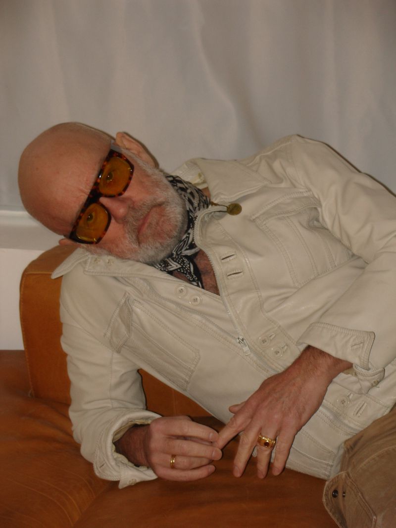 Mature adult male with glasses and a patterned scarf reclines on a brown sofa wearing a light-colored jacket and tan pants.