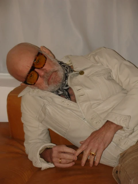 Mature adult male with glasses and a patterned scarf reclines on a brown sofa wearing a light-colored jacket and tan pants.