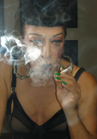 Close-up of a person with dark hair and green nail polish smoking a cigarette, with smoke billowing around their face.