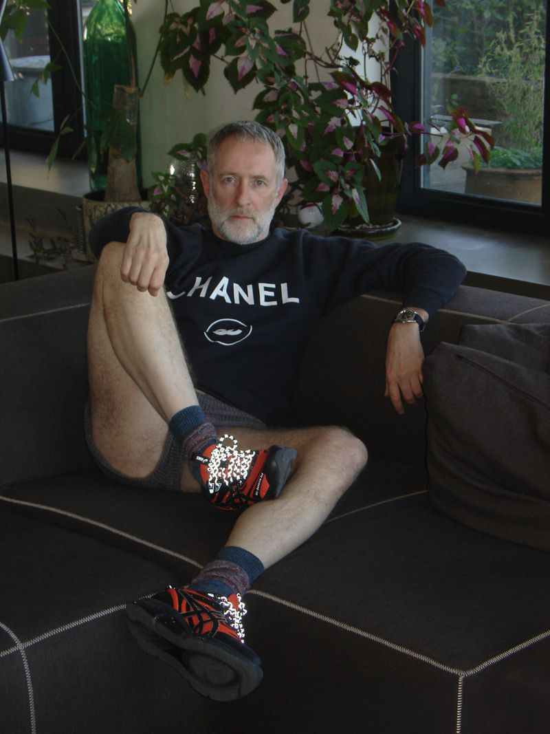 Older man sits on a dark sofa in a sweater and athletic shoes in an indoor environment with plants in the background.
