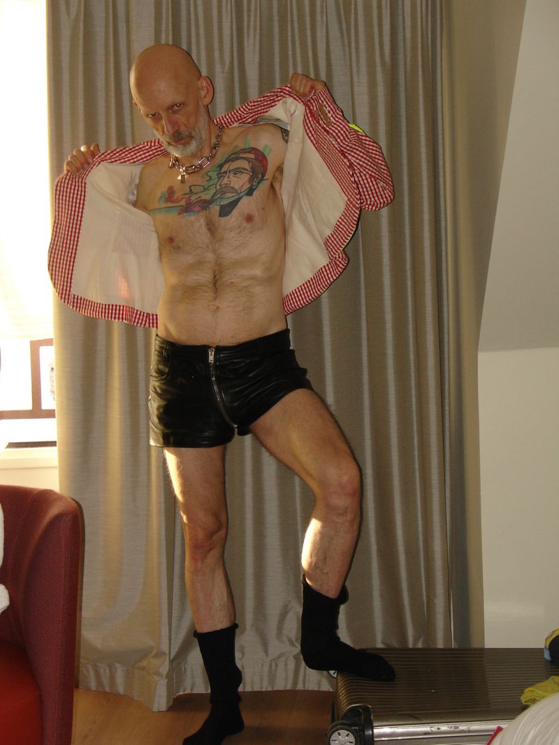 Bald adult male stands on metal step stool, wearing black shorts, socks, patterned jacket, and chain necklace, indoors with neutral drapes.
