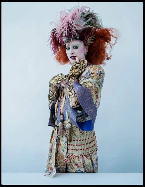 Elegant woman with red hair wearing a feathered hat and patterned robe in a bright photo studio. Full body portrait in a light setting.