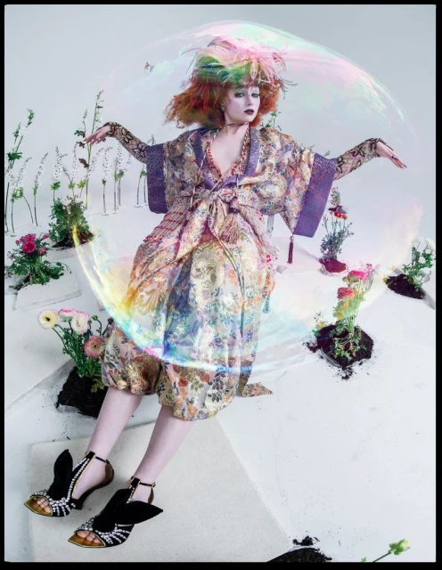 A woman in a floral kimono sits on stairs surrounded by greenery, accentuated by a large bubble and whimsical floral elements.