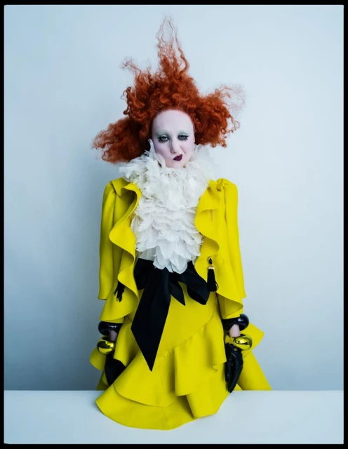 Fashion portrait of a person with pale skin, red hair, and bold makeup wearing a yellow ruffled outfit with a large white collar.