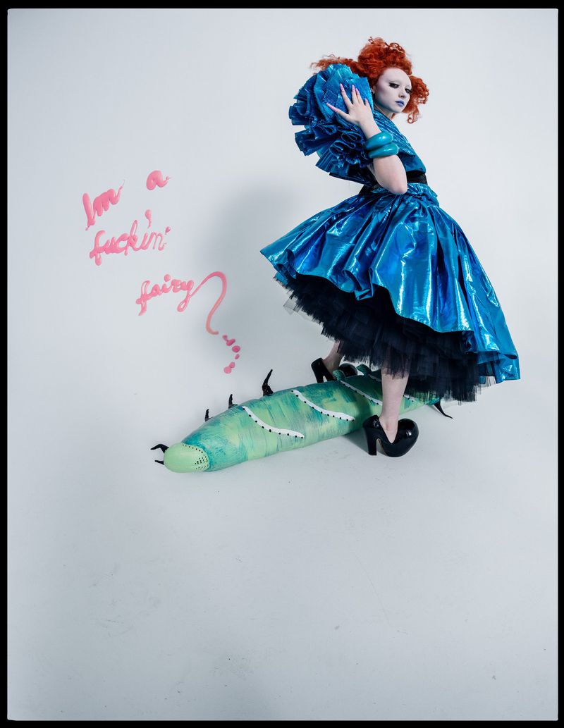 A model in an elaborate blue dress and makeup, standing on a prop with text on the wall in a brightly lit indoor photo studio.