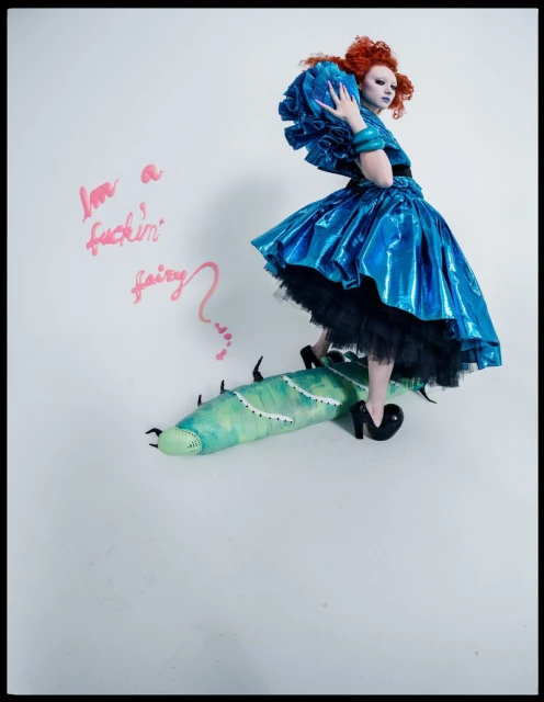 A model in an elaborate blue dress and makeup, standing on a prop with text on the wall in a brightly lit indoor photo studio.