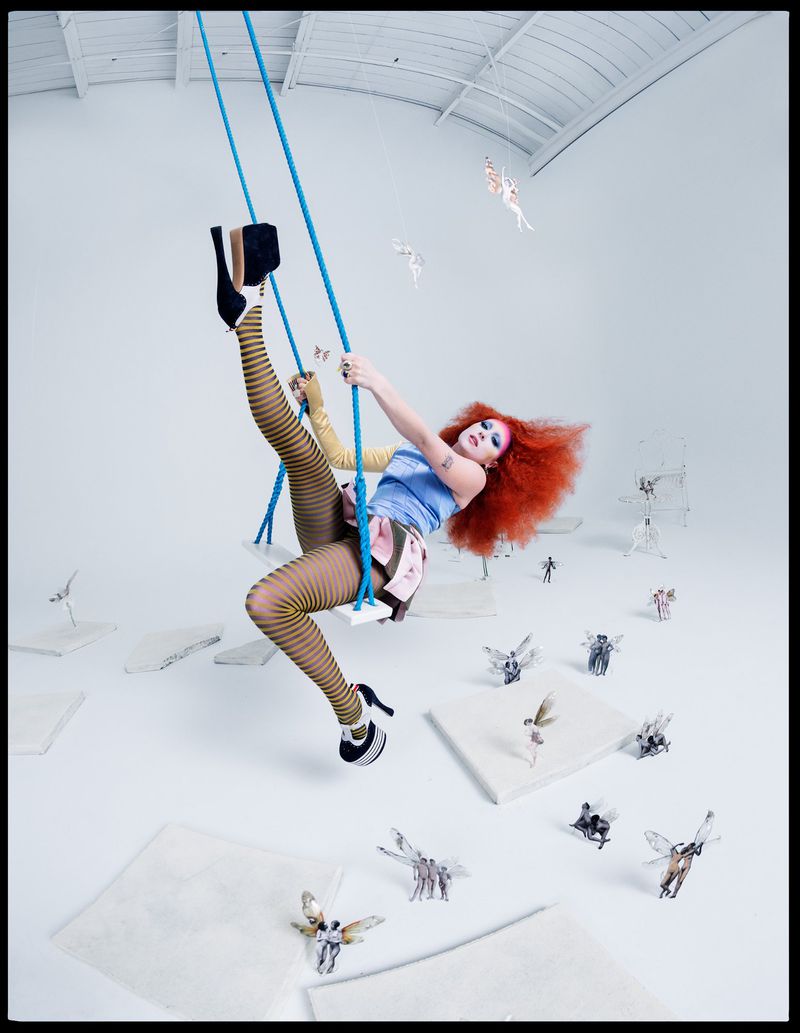 Woman with red hair on swing in a studio setting, wearing striped tights, and high heels. Small fairy-like figures scattered on the floor.