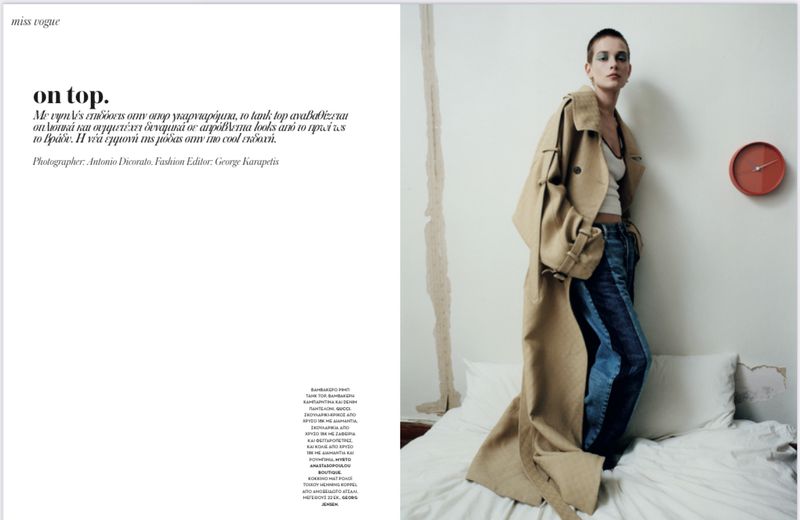 Fashion editorial shot of a woman in a tan trench coat and jeans, standing in a bedroom with white bedding, with a clock on the wall.