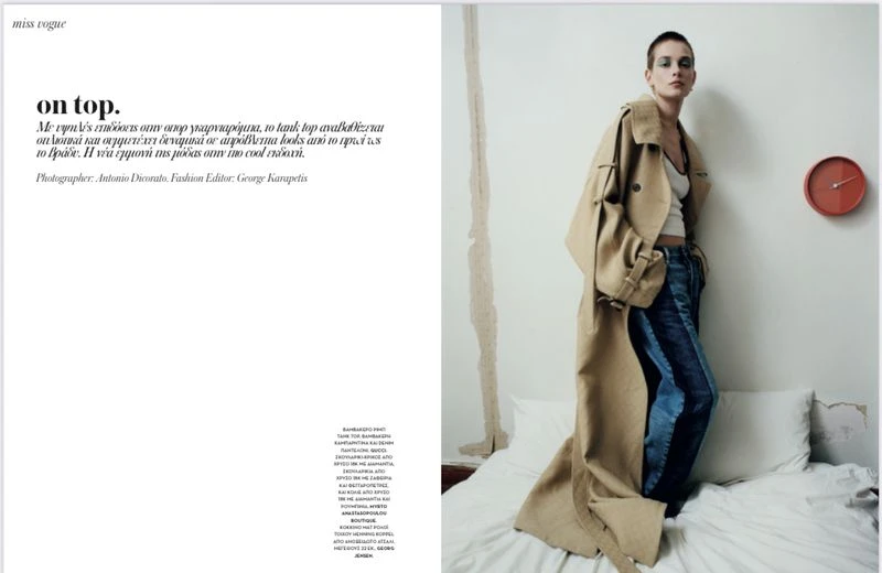 Fashion editorial shot of a woman in a tan trench coat and jeans, standing in a bedroom with white bedding, with a clock on the wall.