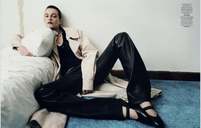 An adult female model is seen indoors wearing a neutral trench coat and black leather pants while reclining against a white wall.