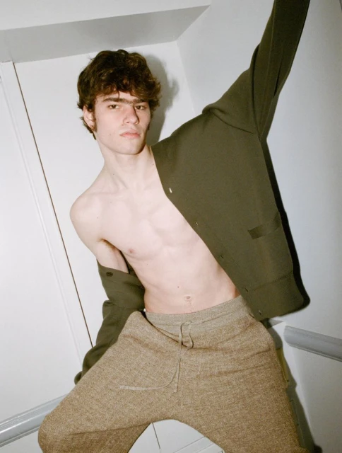 Young man with bare torso poses in front of a white wall wearing grey sweatpants and an olive green jacket, looking at the camera.