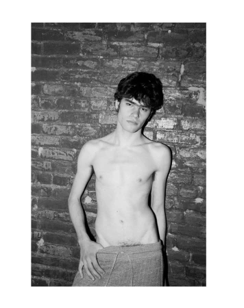 Black and white portrait of a bare-chested young adult male standing against a textured brick wall and wearing a towel wrap.