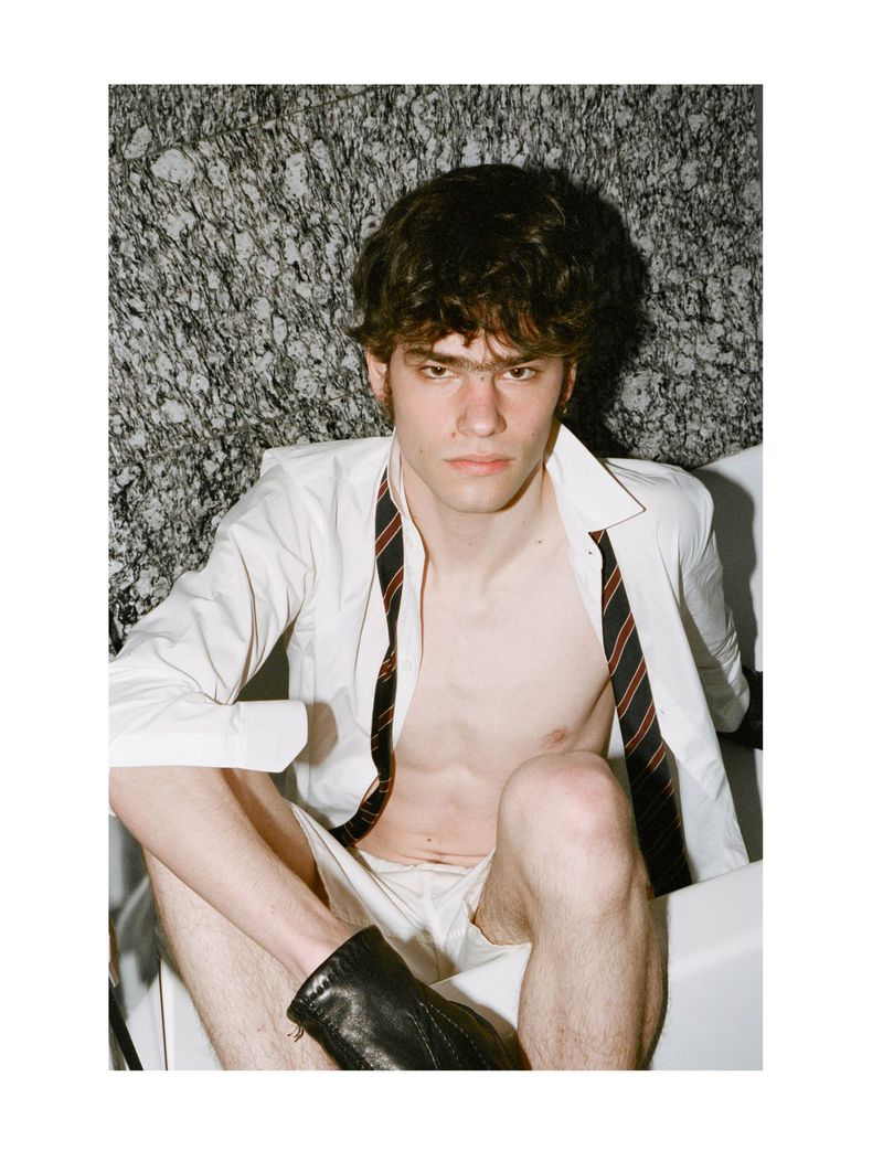 An adult male poses in a bathtub wearing a white shirt, a tie, white briefs and black boots. The background is a marble wall.