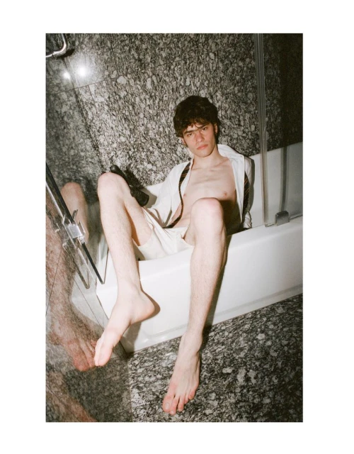 A young adult male sits in a white bathtub, wearing an open white shirt and light shorts, posing in a bathroom with marble walls.