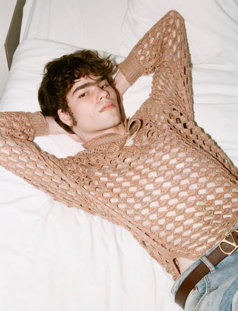 A young adult male poses while laying on a bed wearing a crochet-style shirt with a brown belt, indoors, and looking at the camera.