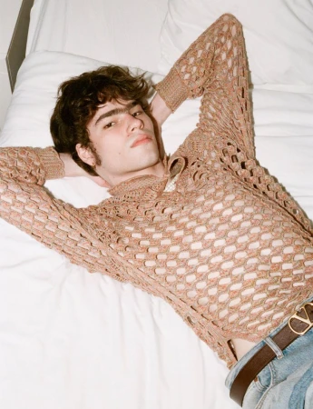 A young adult male poses while laying on a bed wearing a crochet-style shirt with a brown belt, indoors, and looking at the camera.