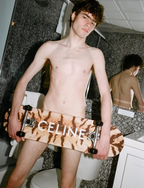 Young adult holding a Celine skateboard in a bathroom with marble walls, reflected in a mirror. Nude male and interior shot.