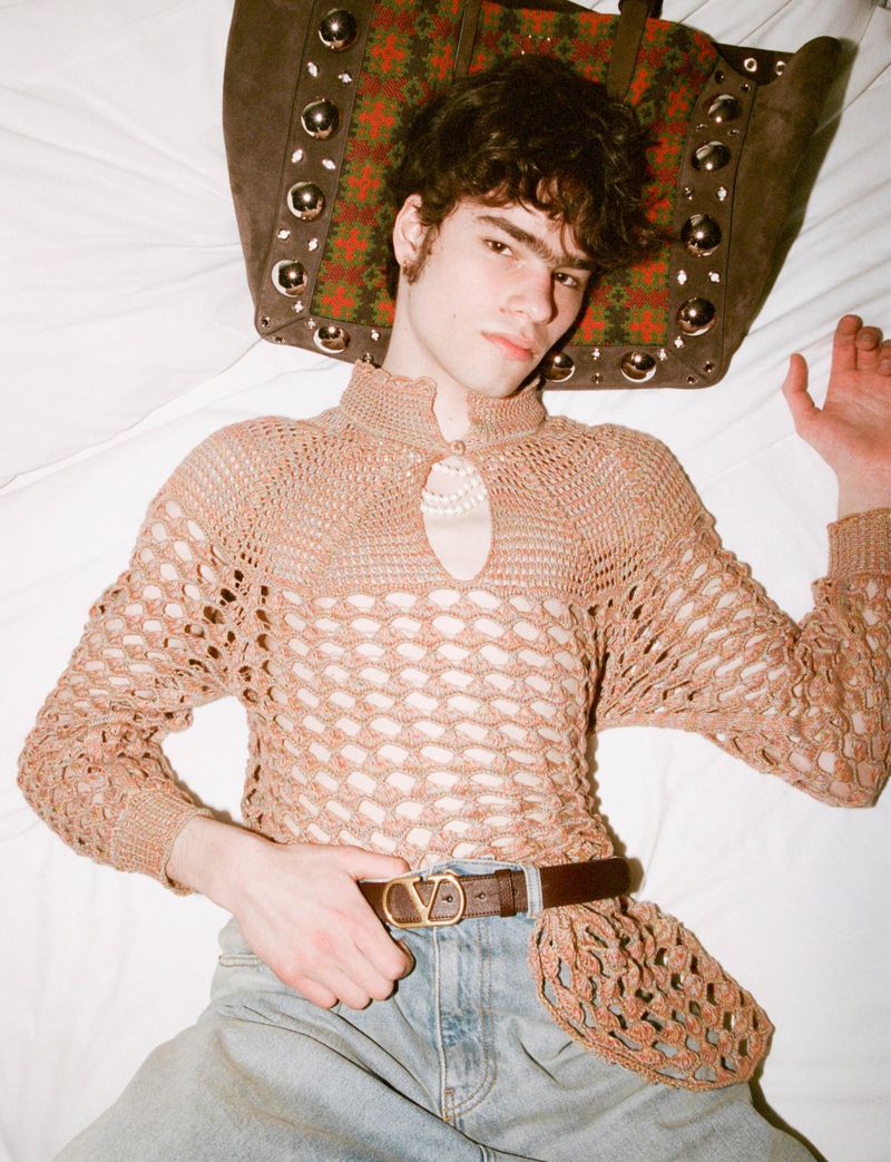 Young adult man lies on a white bed wearing a crocheted top, jeans, and a belt, with a large brown studded bag behind his head.