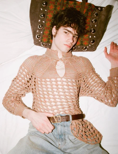 Young adult man lies on a white bed wearing a crocheted top, jeans, and a belt, with a large brown studded bag behind his head.