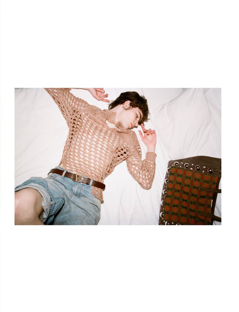 Young adult male lying on a white bed wearing a beige knitted top and denim shorts with a brown belt, next to a patterned brown bag.