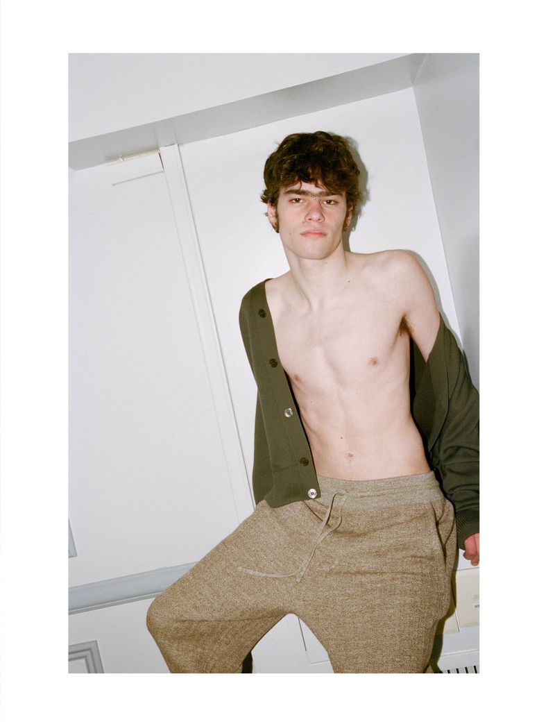 A male model poses indoors wearing a dark green cardigan and tan sweatpants, showcasing a casual yet stylish look against a plain white wall.