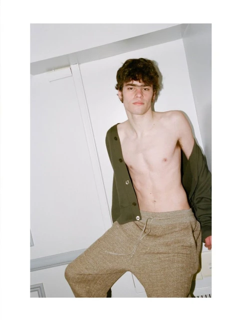 A male model poses indoors wearing a dark green cardigan and tan sweatpants, showcasing a casual yet stylish look against a plain white wall.