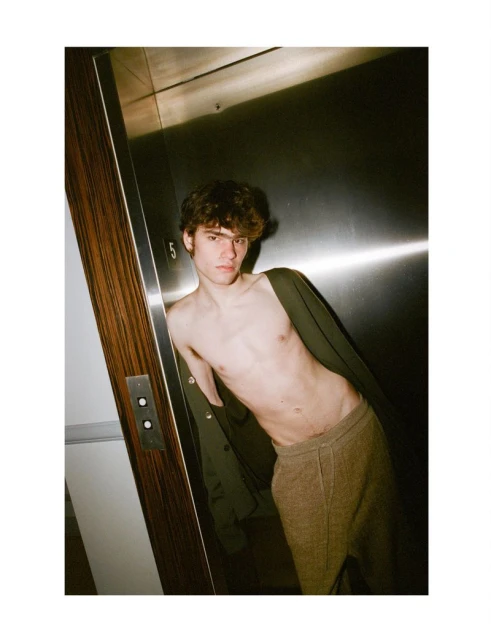 Young adult man with bare torso leans against a silver elevator wearing sweatpants and a green jacket in an indoor setting.